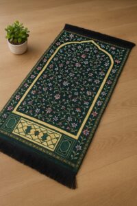 "Elegant Islamic prayer mat (jayenamaz) with floral design on dark green velvet fabric, bordered with intricate yellow and red patterns, placed on wooden floor beside a small indoor plant — perfect for daily salah and spiritual ambiance."