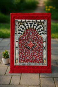 Luxurious red Islamic prayer mat with Ottoman-style arch, floral embroidery, and velvet texture, displayed upright on a stone path with a potted plant.